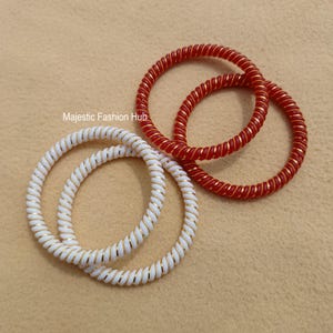 May include: Four spiral bangles are shown: two white with gold detailing and two red with gold detailing, set against a neutral backdrop. The text "Majestic Fashion Hub" is visible.