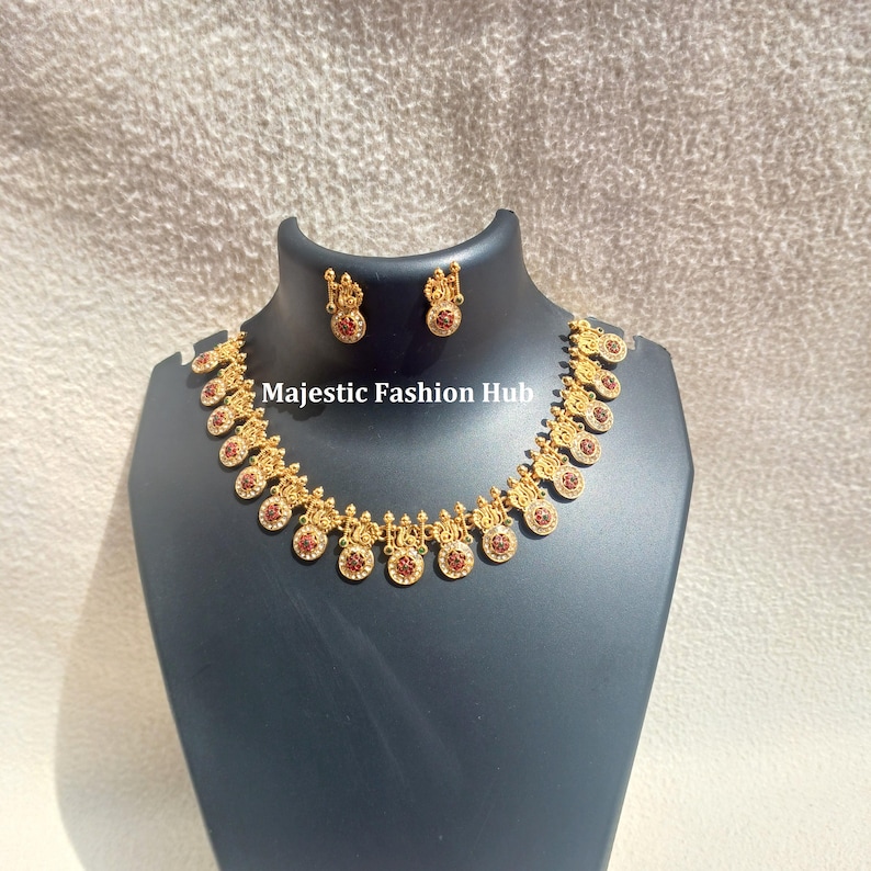May include: Gold-coloured necklace and matching earrings set with red gemstone accents. The necklace features ornate, circular pendants. The text "Majestic Fashion Hub" is visible on the display stand.