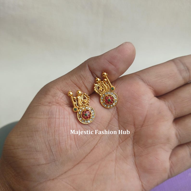 May include: A pair of gold earrings with intricate detailing. Each earring has a circular centrepiece with red and green accents, encircled by clear stones. The earrings have a decorative top section. The text "Majestic Fashion Hub" is visible.