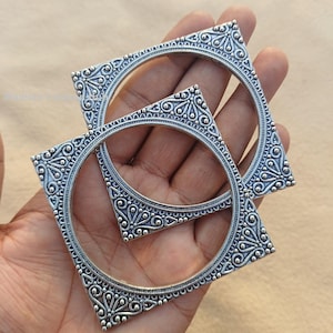 May include: Two silver-colored square bangles with intricate floral and swirl patterns. The bangles are held in a hand, showcasing the detailed design and the contrast between the metal and the skin tone. The jewelry is likely for fashion or cultural wear.