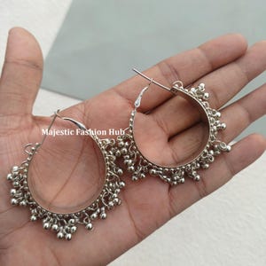 May include: Silver-toned hoop earrings with a beaded design. The earrings are held in a hand, showcasing their circular shape and intricate detailing. The text "Majestic Fashion Hub" is visible.