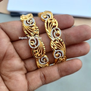 May include: Two gold-colored bangles with intricate filigree designs. The bangles feature a repeating pattern of swirling shapes and floral accents, with silver-colored detailing. The text "Majestic Fashion Hub" is visible.