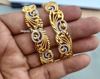 Könnte beinhalten: Zwei goldfarbene Armreifen mit aufwendigen Filigranmustern. Die Armreifen weisen ein sich wiederholendes Muster aus wirbelnden Formen und floralen Akzenten mit silberfarbenen Details auf. Der Text "Majestic Fashion Hub" ist sichtbar.