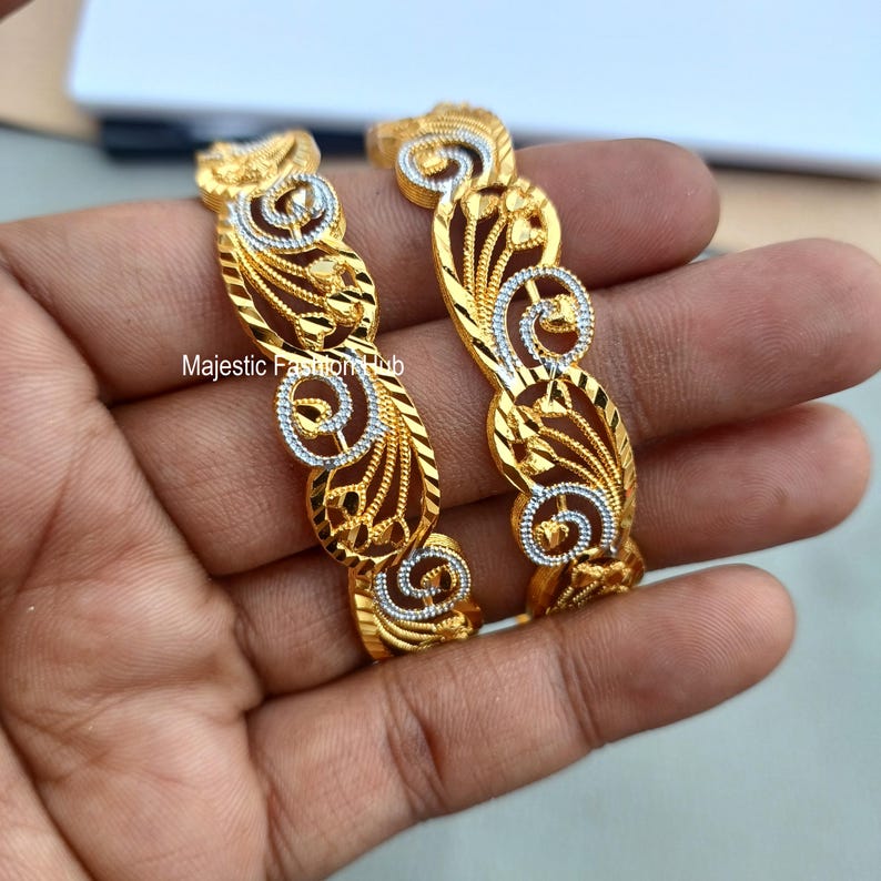 May include: Two gold-colored bangles with intricate filigree designs. The bangles feature a repeating pattern of swirling shapes and floral accents, with silver-colored detailing. The text "Majestic Fashion Hub" is visible.