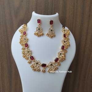 Puede incluir: Un conjunto de collar y pendientes dorados con piedras preciosas rojas y verdes. El collar presenta una serie de piedras rectangulares rojas y detalles florales. Los pendientes reflejan el diseño del collar. Las joyas se exhiben en un maniquí blanco.