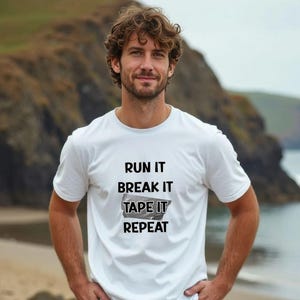 Könnte beinhalten: Weißes T-Shirt mit Rundhalsausschnitt und dem Text "RUN IT BREAK IT TAPE IT REPEAT" in Schwarz. Das T-Shirt wird von einer Person mit lockigem braunem Haar getragen, die vor einem verschwommenen Hintergrund im Freien steht.