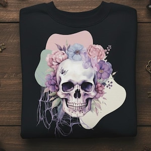 Könnte beinhalten: Schwarzes Sweatshirt mit Rundhalsausschnitt und einem Totenkopf-Design, verziert mit lila, rosa und blauen Blumen. Eine Spinne befindet sich auf dem Schädel. Das Design ist vor abstrakten Formen platziert. Ein gruseliger und stilvoller Artikel.