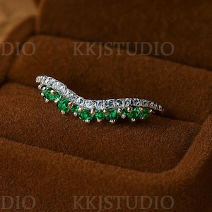 Pode incluir: Um anel curvo com uma fileira de pedras preciosas verdes e pequenas pedras transparentes, em uma banda de prata. O anel é exibido em uma caixa de anel de veludo marrom aberta. O fundo é de cor bege neutra.