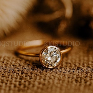 May include: A gold ring featuring a large, round, clear gemstone. The ring rests on a textured, brown surface. The ring's band is slender, and the gemstone is the central element.