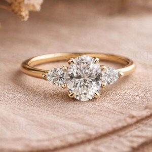 May include: A gold engagement ring featuring an oval-cut diamond as the central stone, flanked by two pear-shaped diamonds. The ring has a slender gold band. The diamonds are set to catch the light.