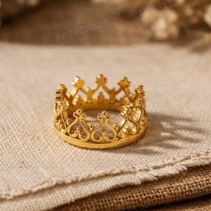 Puede incluir: Un anillo de corona dorado con detalles intrincados. El anillo presenta una serie de arcos que apuntan hacia arriba y elementos decorativos, que se asemejan a una corona en miniatura. El anillo se muestra sobre una tela texturizada de tono neutro.
