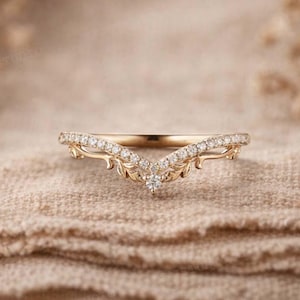 Op de afbeelding: Een goudkleurige ring met een V-vormig ontwerp. De ring is versierd met kleine, heldere edelstenen en bladvormige details. De ring is geplaatst tegen een neutrale, getextureerde achtergrond. Waarschijnlijk een trouw- of verlovingsring.