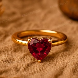 Puede incluir: Un anillo de oro con una piedra preciosa roja oscura en forma de corazón. La banda del anillo es lisa y redondeada, y la piedra preciosa está engastada con pequeñas garras doradas. El anillo se muestra sobre una superficie texturizada y marrón.