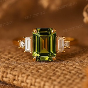 Puede incluir: Un anillo de oro con una gran piedra preciosa verde oliva con corte esmeralda como pieza central, flanqueada por dos piedras preciosas rectangulares y transparentes. Pequeñas piedras preciosas redondas y transparentes están engastadas a ambos lados de las piedras rectangulares. El anillo está sobre una superficie marrón texturizada.