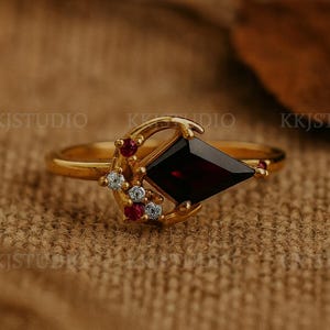 May include: A gold ring featuring a dark red, kite-shaped gemstone as the centerpiece. The ring is accented with small red and white gemstones, set in a crescent moon design. The ring is displayed on a textured, brown surface.