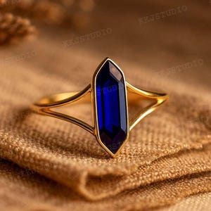 May include: A gold ring featuring a vibrant, elongated, deep blue gemstone. The ring's band splits into a delicate design on either side of the gemstone, creating an elegant and modern aesthetic. The ring is set against a textured, tan fabric.