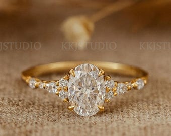 May include: A gold engagement ring featuring an oval-cut central gemstone, surrounded by smaller accent stones. The ring has a slender band. The design is classic and elegant, set against a neutral backdrop. The ring is a symbol of commitment.
