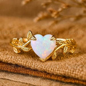 May include: A gold ring with a heart-shaped opal gemstone. The ring has a delicate leaf design on the band. The opal is a milky white colour and the ring is set on a brown surface.