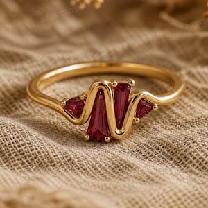 May include: A gold ring with a unique design. The ring features three red gemstones, two triangular and one rectangular, set in a wavy pattern. The ring is displayed on a textured, neutral-colored surface.