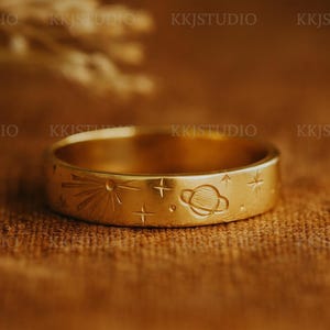 Könnte beinhalten: Ein goldfarbener Ring mit eingravierten Himmelsmotiven, darunter ein Sonnenstrahl, Sterne und ein Planet. Der Ring ist vor einem strukturierten, braunen Hintergrund platziert, der die filigranen Details des Schmucks im Weltraum-Design hervorhebt.