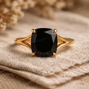 Puede incluir: Un anillo de oro con una gran piedra preciosa negra de corte cuadrado. El anillo tiene un diseño de caña dividida y está colocado sobre una tela de tono neutro. El anillo es una joya.