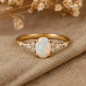 May include: A gold ring featuring a central oval-shaped opal gemstone held by four prongs. The band is accented on both sides by a cluster of three small round clear stones and one pear-shaped clear stone, all set in a polished gold metal band.