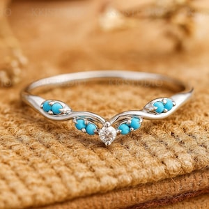 Op de afbeelding: Een zilveren ring met een gebogen ontwerp, met kleine turkooizen stenen en een centrale diamant. De ring is geplaatst tegen een achtergrond van getextureerde, bruinachtige stof, die de details van de sieraden laat zien.