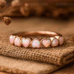 Könnte beinhalten: Ein roségoldener Ring mit herzförmigen Opalsteinen. Die Opale haben eine perlmuttartige, irisierende Qualität und sind in einem zarten Band gefasst. Der Ring wird auf einer strukturierten, neutral getönten Oberfläche gezeigt. Der Text "KKJSTUDIO" ist im Hintergrund sichtbar.