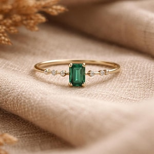 Puede incluir: Un anillo de oro con una esmeralda rectangular en el centro. Pequeños diamantes redondos están engastados a ambos lados de la esmeralda. El anillo está sobre una tela de color neutro.