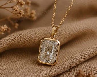Peut inclure: Un collier pendentif en or avec une grande pierre précieuse rectangulaire et transparente. La pierre est sertie dans un cadre en or et suspendue à une fine chaîne en or. Le collier est présenté sur un tissu beige texturé.