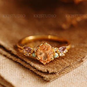 Könnte beinhalten: Ein goldener Ring mit einem ovalen, pfirsichfarbenen Edelstein als Mittelpunkt. Der Ring ist mit kleineren lila, türkisfarbenen und klaren Edelsteinen verziert. Der Ring liegt auf einer strukturierten braunen Oberfläche. Der Markenname "KKJSTUDIO" ist sichtbar.