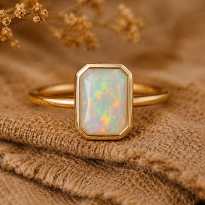 May include: A gold ring featuring a rectangular, faceted opal gemstone set in a smooth bezel setting. The opal displays iridescent flashes of green, orange, and yellow colors against a milky white base. The band is a simple, polished gold metal.