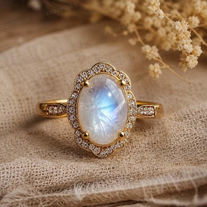 May include: A gold ring featuring an oval moonstone with an iridescent sheen, encircled by small, clear gemstones. The ring's band is gold, with additional small stones on either side of the central moonstone. The ring is set on a beige fabric.