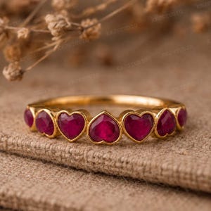 May include: A gold ring featuring heart-shaped ruby gemstones. The ring has a band of gold and is set with multiple heart-cut rubies. The gemstones are a deep red color, and the ring is displayed on a textured surface.