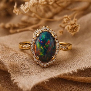 May include: A gold ring featuring an oval black opal gemstone with vibrant flashes of color. The opal is surrounded by a halo of small, clear gemstones. The ring's band is also adorned with small gemstones.