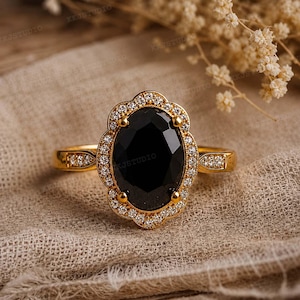 Op de afbeelding: Een gouden ring met een grote, ovaal geslepen zwarte steen in het midden, omringd door een geschulpte halo van kleine, heldere pavé-gezette steentjes. De ringband is aan beide zijden ook versierd met kleine heldere steentjes voor detail.