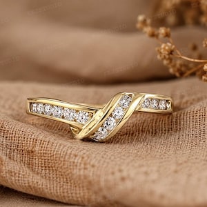 May include: A gold-colored ring with a curved design, set with multiple small, clear gemstones. The ring is on a tan fabric surface, showcasing its intricate details and elegant style. The ring's design is a V-shape.