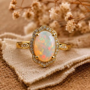 Op de afbeelding: Een gouden ring met een ovale opaal edelsteen, omringd door kleine diamanten. De opaal vertoont kleurspel met rode, oranje en groene flitsen. De ringband is ook versierd met kleine diamanten.