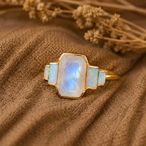 May include: A gold ring featuring a large, rectangular, translucent moonstone centre stone with a blue sheen, flanked by two smaller, rectangular, iridescent opal side stones on each side, set in a polished gold metal band.