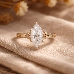 May include: A gold engagement ring with a marquise-cut diamond. The ring has detailed filigree work on the band and around the diamond setting. The ring is displayed on a neutral-toned fabric.