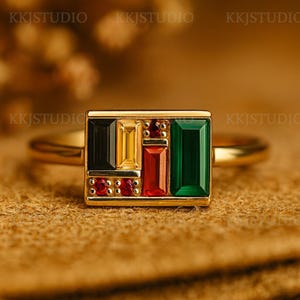 May include: A gold ring featuring a rectangular design with various colored gemstones. The gemstones include black, yellow, red, and green, arranged in a geometric pattern. Small red stones are also set along the bottom edge of the rectangle.