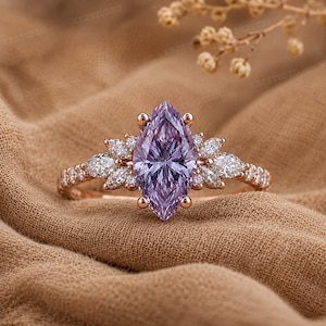 May include: A rose gold ring featuring a large marquise-cut lavender centre stone flanked by clusters of smaller marquise-cut white stones on each side. The band is thin and metallic, showcasing a delicate design with prong settings for each stone.