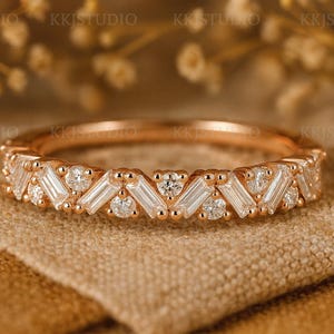 Puede incluir: Anillo de oro rosa con diamantes de talla baguette y redondos alternados. El anillo presenta un patrón repetitivo de piedras rectangulares y circulares, engastadas en una banda delicada. El anillo se muestra sobre una tela beige.