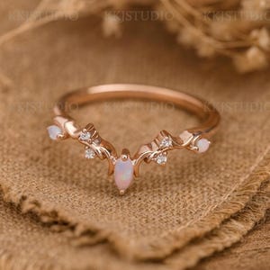 Puede incluir: Anillo de oro rosa con una piedra central de ópalo en forma de marquesa y ópalos más pequeños a cada lado. El anillo presenta un diseño delicado y curvado con pequeñas piedras de acento brillantes. El anillo está sobre una superficie texturizada de tono neutro.