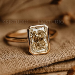 Può includere: Un anello d'oro con una grande pietra preziosa rettangolare giallo chiaro. La pietra preziosa è incastonata in una lunetta d'oro. L'anello è su un tessuto beige strutturato. È un gioiello.