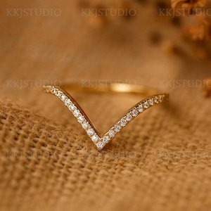 Może przedstawiać: Złoty pierścionek w kształcie litery V z małymi, przezroczystymi kamieniami szlachetnymi wzdłuż górnych krawędzi. Pierścionek jest prezentowany na teksturowanej, beżowej powierzchni. Nazwa marki "KKJSTUDIO" jest widoczna w tle.