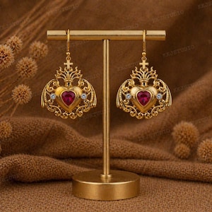 May include: Gold-toned heart-shaped earrings with a red center stone and small clear accent stones. The earrings have a decorative filigree design and a cross at the top. They are displayed on a gold jewelry stand.