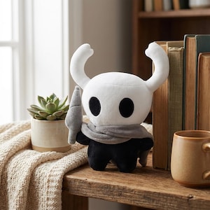 以下が含まれることがあります： 白い頭、大きな黒い目、湾曲した角を持つキャラクターのぬいぐるみ。キャラクターは灰色のスカーフを着用し、灰色の剣を持っています。体は黒です。おもちゃは、多肉植物とマグカップのある木製の棚にあります。