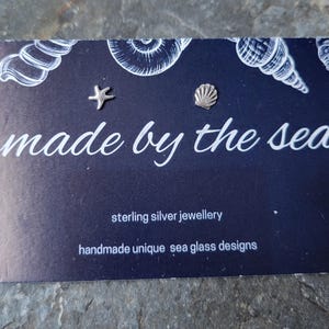 May include: A dark blue business card with the text "made by the sea" in white script. The card features seashell illustrations and two small silver charms: a starfish and a scallop shell. The card also reads "sterling silver jewellery".