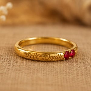 May include: A gold band ring featuring the engraved script text Till Death and two small round red gemstones set side by side. The polished metal surface reflects light, highlighting the cursive lettering and the vibrant color of the stones.